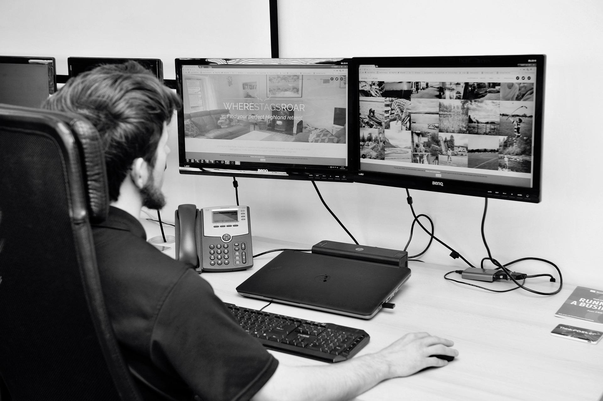 Bir çalışanın masa başında iki bilgisayar ekranında web sayfalarını inceleyerek çalıştığı ofis ortamı. Masada klavye, dizüstü bilgisayar ve ofis telefonu bulunuyor.